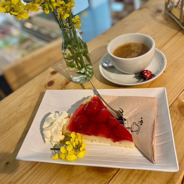 Erdbeerkuchen, Café Fehmarn Käsekuchen mit Erdbeeren, Sahne, gelben Blumen und einer Tasse Kaffee auf einem Holztablett.